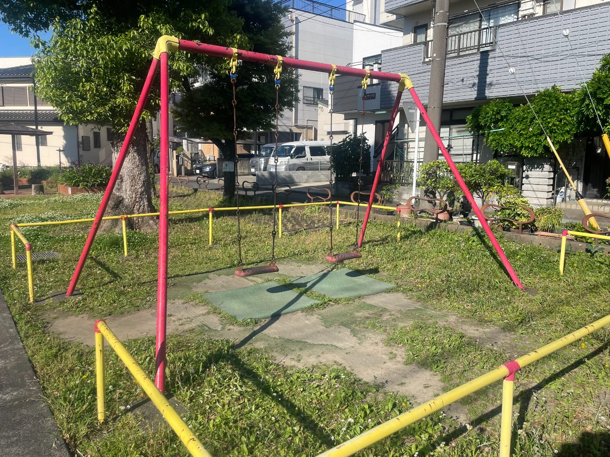 後谷第一公園の赤いブランコ2連。黄色のフェンスに囲まれ緑の草地に設置されている