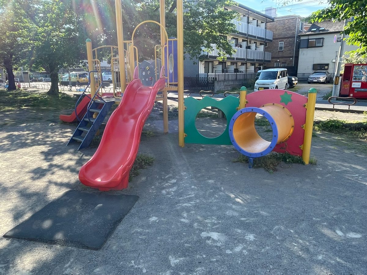 赤いすべり台とカラフルなトンネル付き複合遊具。木陰の中に設置