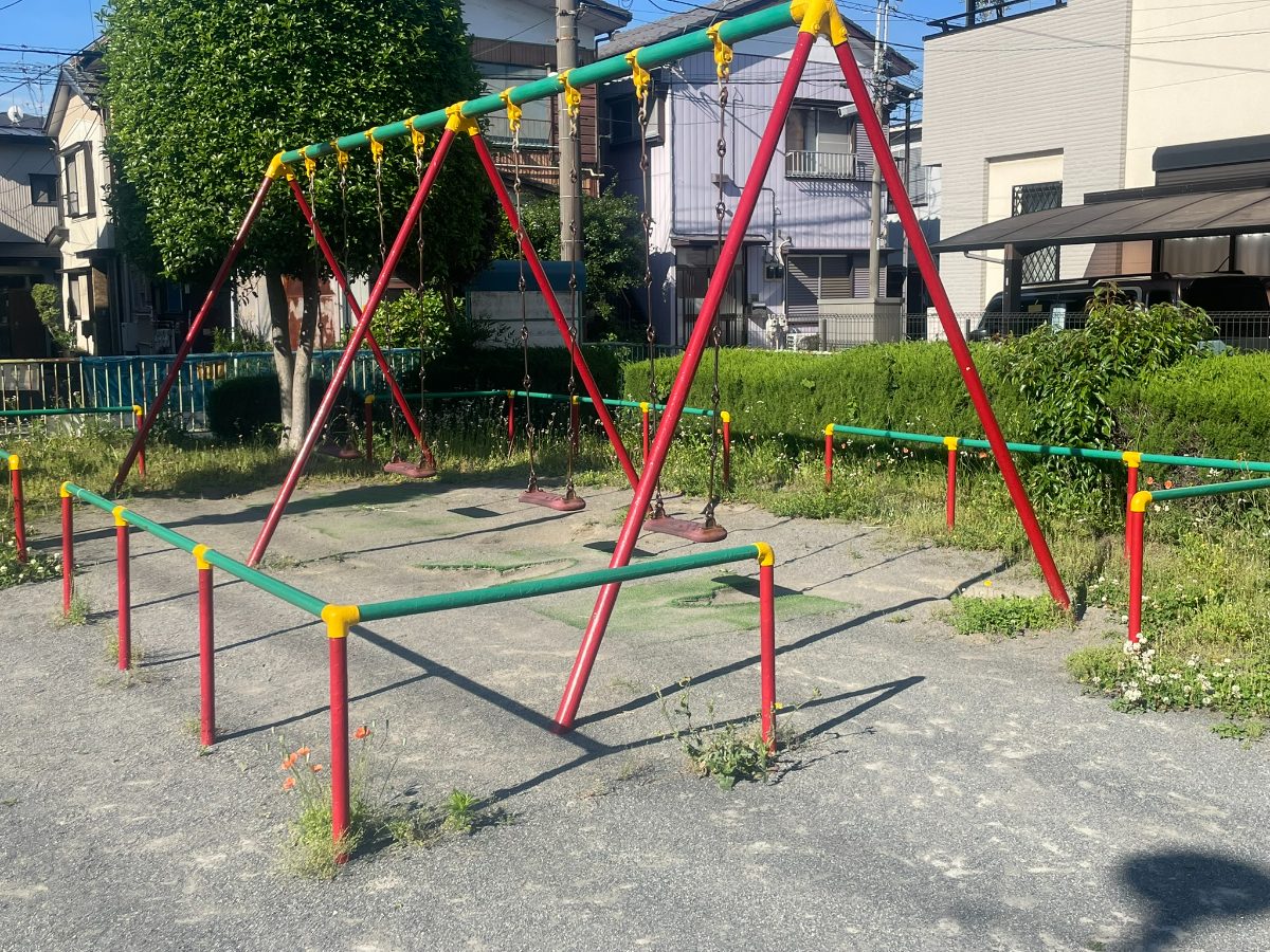赤と緑のブランコ2連。青空と緑の生垣を背景に設置されている