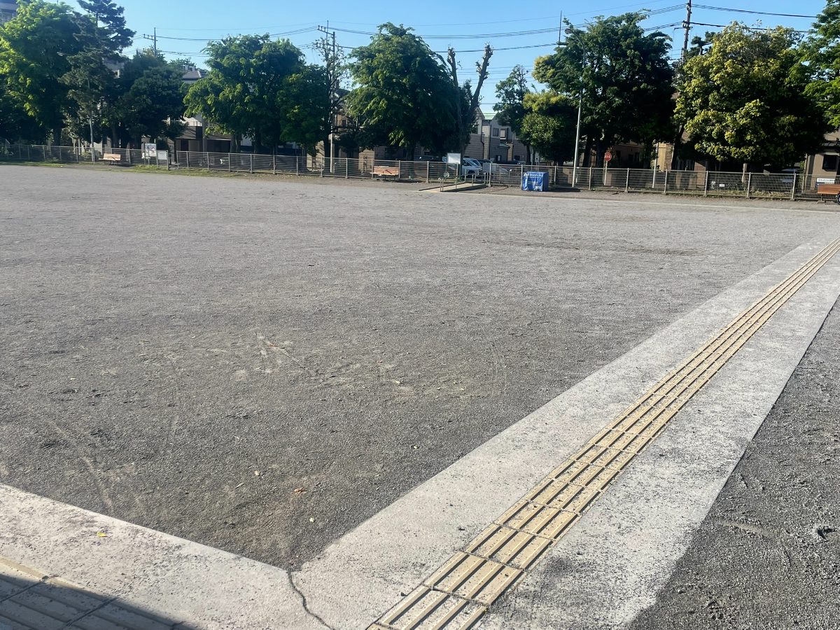 中町多目的広場の広大な砂地グラウンド全景。奥に並木と青空が広がる